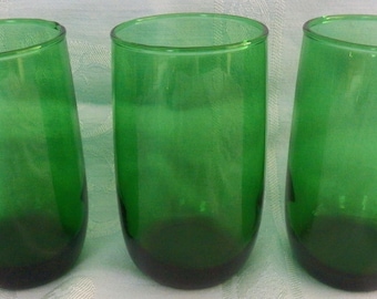 May include: Three green-tinted drinking glasses. The glasses are cylindrical with a rounded base and a slightly tilted shape. The color transitions from a darker green at the base to a lighter green towards the top. The glasses are empty.