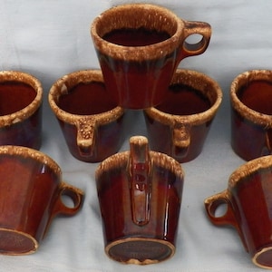 May include: A set of seven ceramic mugs featuring a deep brown glossy glaze with a textured, lighter tan drip pattern around the upper rim and handle. Each mug has a sturdy side handle and a tapered cylindrical body with a smooth finish.