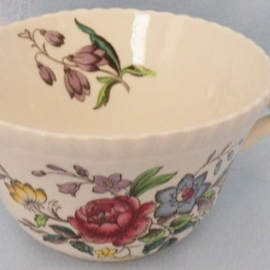 May include: Cream-colored teacup with a scalloped rim and a floral design. The cup features a handle and is decorated with purple, red, yellow, and blue flowers and green leaves. A delicate piece for tea or coffee.