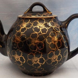 May include: A round ceramic teapot featuring a glossy black finish with a repeating gold floral pattern. The teapot includes a matching lid with a curved handle, a curved spout, and a large side handle, all accented with gold trim along the edges.