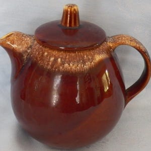 May include: A brown ceramic teapot with a lid and handle. The teapot has a glossy finish and a two-tone design, with a lighter brown band around the rim. The lid has a small knob on top. The teapot is designed for serving tea.