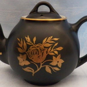 May include: A matte black ceramic teapot featuring a gold floral design on the front. The teapot has a curved handle, a spout with a gold accent line, and a matching lid with a gold rim. The gold pattern depicts a central rose with leaves.