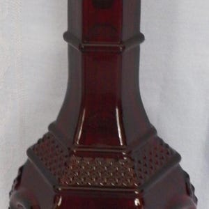 May include: A deep red glass decanter featuring a hexagonal body with intricate embossed diamond patterns and circular motifs. The decanter includes a matching removable stopper with a textured, faceted diamond-shaped top and a tall neck.
