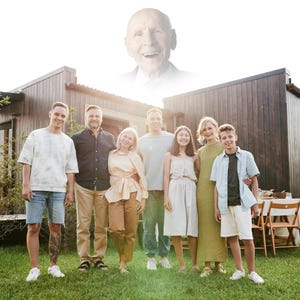 Op de afbeelding: Een groep mensen, waaronder een lachende oudere man op de achtergrond, poseert voor een houten huis. De groep bestaat uit mannen, vrouwen en kinderen, gekleed in vrijetijdskleding, staand op groen gras.