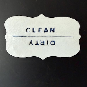 Puede incluir: Un letrero de cerámica blanca con las palabras "CLEAN" y "DIRTY" en letras azul oscuro. El letrero tiene un borde decorativo festoneado y una línea azul oscuro que separa las dos palabras. El fondo es negro.