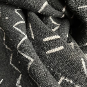 May include: Close-up of a dark gray textile with a white geometric pattern. The fabric is folded, showing texture and depth. The design features zig-zag and linear shapes, creating a modern, abstract look. The textile appears to be woven.