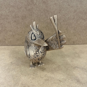 May include: A silver-coloured bird figurine with intricate floral patterns. The bird is holding two sticks in its wing. The figurine has a detailed beak, eyes, and feet. The overall design is ornate and decorative.