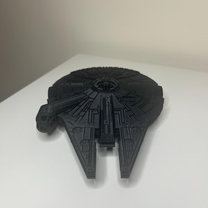 May include: A black model of the Millennium Falcon starship, featuring detailed paneling and design. The model has a distinct cockpit and iconic shape. The starship is a popular item from the Star Wars franchise.