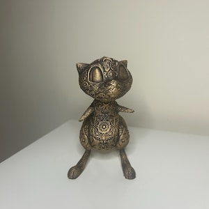 May include: A bronze cat figurine with detailed floral and swirl patterns. The cat has large, round eyes and is posed in a seated position with its arms outstretched. The sculpture has a detailed, textured surface.