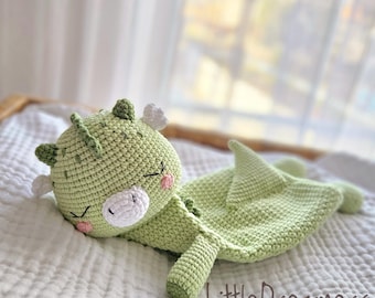 May include: A green crocheted dragon stuffed animal with a white face and pink cheeks. The dragon is lying on its side with its head resting on a white blanket. The text "Little Dreamers" is visible in the bottom right corner of the image.