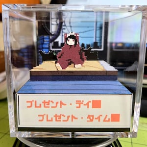 May include: A pixelated diorama in a clear acrylic box. It features a figure in a red outfit with a panda-like hood, seated on a platform. The diorama includes a cityscape backdrop and Japanese text that reads "Present Day" and "Present Time".