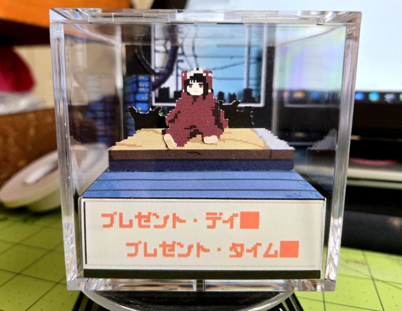 May include: A pixelated diorama in a clear acrylic box. It features a figure in a red outfit with a panda-like hood, seated on a platform. The diorama includes a cityscape backdrop and Japanese text that reads "Present Day" and "Present Time".