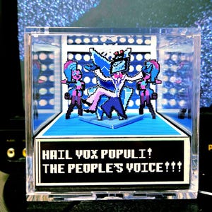 Puede incluir: Un diorama pixelado en una caja de acrílico transparente. La figura central, en azul y blanco, tiene los brazos extendidos y está flanqueada por dos figuras. La base de la caja muestra el texto "HAIL VOX POPULI! THE PEOPLE'S VOICE!!!"