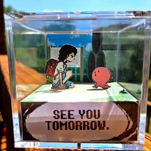 Peut inclure: Diorama dans une boîte en acrylique transparente avec un pixel art d'une personne avec un sac à dos et une pieuvre rose. Le diorama a un fond peint et le texte "SEE YOU TOMORROW." est affiché à l'avant.