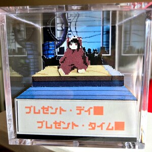 May include: A pixelated diorama in a clear acrylic box. The scene features a figure in a maroon outfit sitting on a platform with a blue and brown base. The background depicts a cityscape. Japanese text is visible on the base.