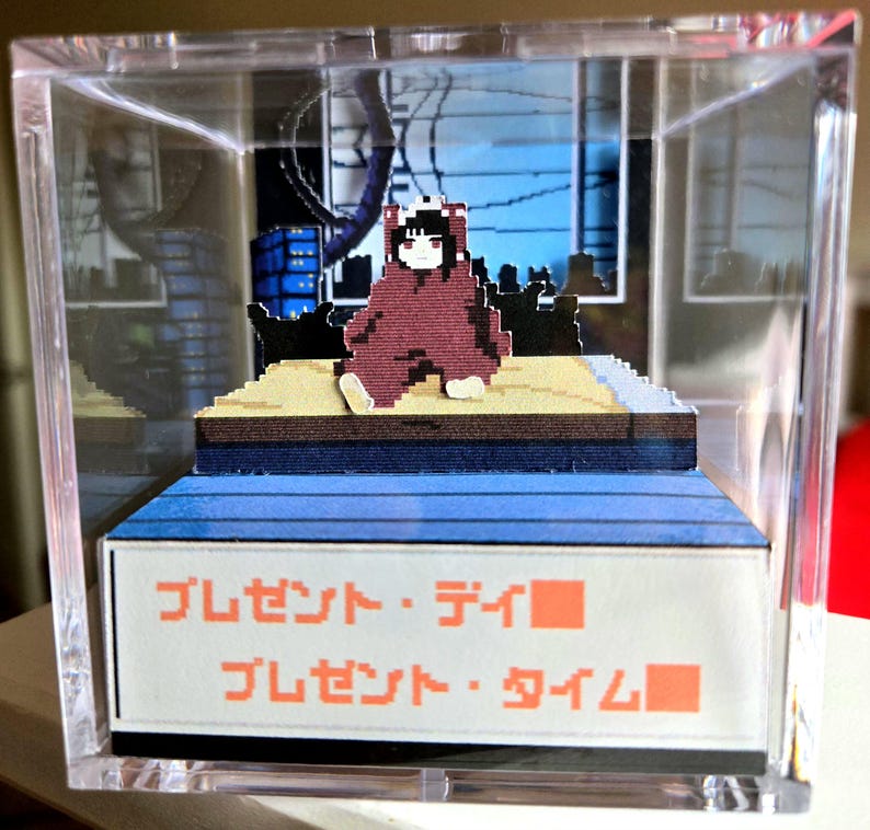 May include: A pixelated diorama in a clear acrylic box. The scene features a figure in a maroon outfit sitting on a platform with a blue and brown base. The background depicts a cityscape. Japanese text is visible on the base.