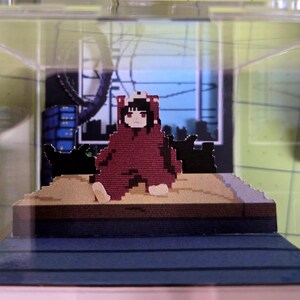 May include: A pixelated figure in a red hooded outfit sits on a platform inside a clear display case. The figure has black hair and red eyes. The background features a pixelated cityscape with blue and black buildings.