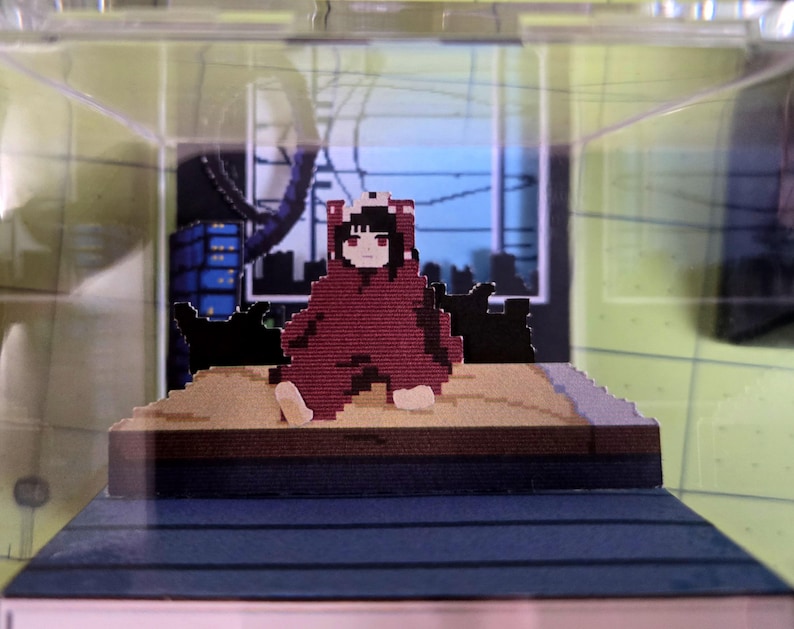 May include: A pixelated figure in a red hooded outfit sits on a platform inside a clear display case. The figure has black hair and red eyes. The background features a pixelated cityscape with blue and black buildings.