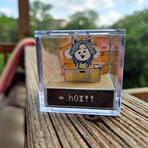 Puede incluir: Una caja de presentación de cubo de plástico transparente con una pequeña figura de papel de un personaje del videojuego "Undertale". El personaje es un monstruo azul y amarillo con una cara blanca y un corazón rojo. El personaje está sentado en un escritorio con una taza de té y un cartel que dice "TEM'S SHOP". La base de la caja de presentación tiene el texto "* HOI!!" impreso en ella.