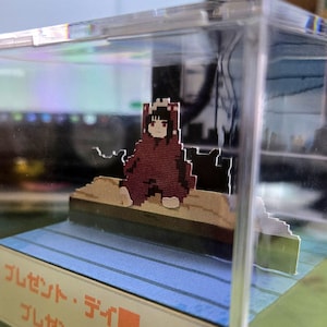May include: A pixelated figure in a red hooded cloak sits on a wooden platform inside a clear acrylic display case. The figure has black hair and a neutral expression. The base of the display case has Japanese text.