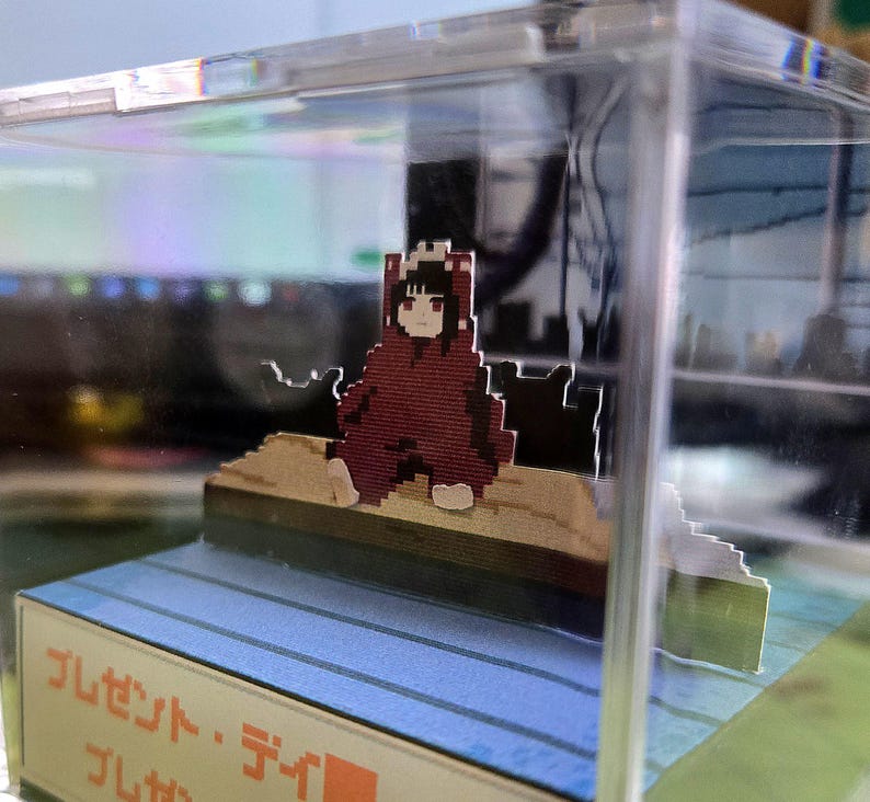 May include: A pixelated figure in a red hooded cloak sits on a wooden platform inside a clear acrylic display case. The figure has black hair and a neutral expression. The base of the display case has Japanese text.