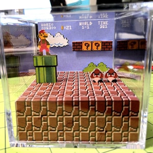 May include: A miniature diorama of a classic video game scene, featuring a pixelated Mario character standing on a green pipe. The scene includes brick platforms, question mark blocks, and Goombas, all encased in a clear acrylic cube. The background displays game details.