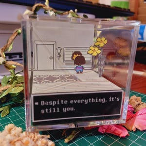 May include: A clear acrylic box diorama featuring a pixelated character from the video game Undertale. The character is standing in a room with a door and a window. The text "Despite everything, it's still you." is displayed on a black background.