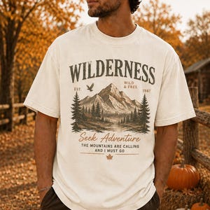 May include: An off-white short-sleeve t-shirt featuring a graphic print of a mountain landscape with pine trees, a cabin, and an eagle. The text reads WILDERNESS, EST. 1987, WILD & FREE, Seek Adventure, THE MOUNTAINS ARE CALLING AND I MUST GO.