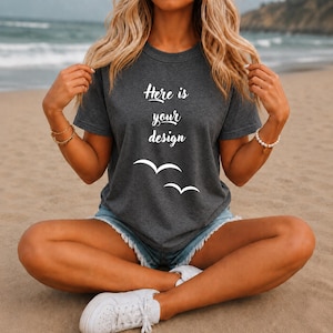 May include: A dark gray t-shirt with the text "Here is your design" and two white bird graphics. The shirt is paired with denim shorts and white sneakers. The person is sitting on a sandy beach.