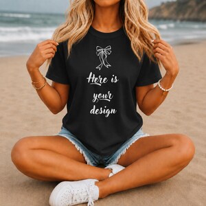 May include: Black t-shirt with the text "Here is your design" in a white script font. The shirt features a white bow graphic above the text. The model is wearing denim shorts and white sneakers while sitting on the beach.