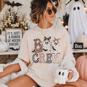 以下が含まれることがあります： BOO CREWという文字がプリントされたクリーム色の半袖Tシャツ。デザインには、リボンをつけた2体のゴースト、ヒョウ柄、チェック柄、そして小さなコウモリや星のイラストが描かれています。