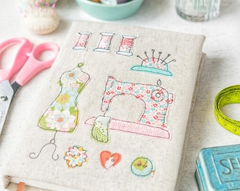 May include: A linen-covered notebook with embroidered sewing-themed designs, including a dress form, sewing machine, and spools of thread. Pink scissors, a pincushion, and a measuring tape are also visible, suggesting a crafting theme.