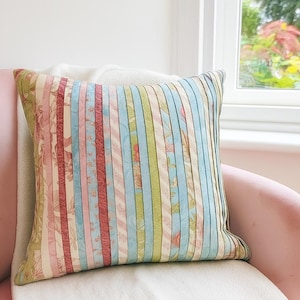 May include: Square cushion with a patchwork design. The cushion features vertical stripes in various colours, including pink, blue, green, and floral patterns. The cushion is placed on a pink chair and a white blanket.