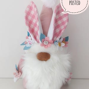 May include: A pink and white gnome with a white beard and pink gingham bunny ears. The gnome is wearing a flower crown with pink, blue, and white flowers. The gnome is standing on a white surface. "Printed and Posted" is written in a speech bubble above the gnome.