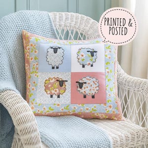 Może przedstawiać: Kwadratowa poduszka dekoracyjna z czterema wzorami owiec na białym wiklinowym krześle. Poduszka ma kwiatową obwódkę i tekst "PRINTED & POSTED" w dymku. Owieczki są haftowane na niebieskich i różowych kwadratach.