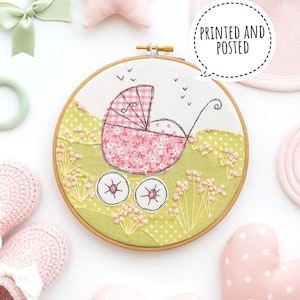 Op de afbeelding: Geborduurde babywagen-hoepelkunst met een roze bloemenontwerp en de tekst "PRINTED AND POSTED". Het kunstwerk wordt omringd door baby-accessoires, waaronder een strik, ster en babyschoentjes.