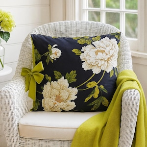 May include: A cushion featuring a dark blue floral design with large white peonies and green leaves. It rests on a white wicker chair, with a lime green bow and a matching throw blanket.