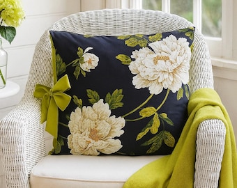 May include: A decorative pillow with a dark blue background and large white peony floral print. The pillow is on a white wicker chair with a lime green bow and a matching throw blanket.