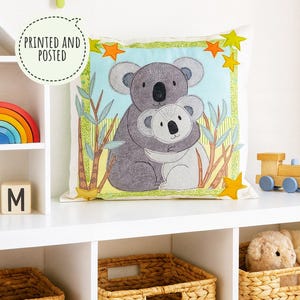 May include: A square cushion with a design of a grey koala embracing a smaller, light grey koala. The image includes green eucalyptus leaves and yellow stars. The cushion is on a white shelf with toys and baskets.