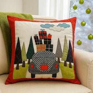 May include: Decorative square pillow with a red border, featuring an applique design of a car carrying gifts on a road through a forest. The car is gray and black, and the gifts are wrapped in plaid and solid colors.