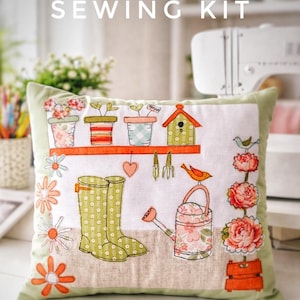 May include: A decorative pillow with a garden-themed embroidery design. The pillow features a white center with images of flower pots, a birdhouse, boots, and a watering can. The text "SEWING KIT" is at the top. The pillow has a green border and orange floral accents.