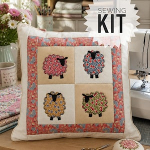 May include: A square fabric cushion featuring a patchwork design with four sheep appliqués in pink, blue, yellow, and green floral patterns, framed by a coral floral border. The text SEWING KIT is printed in black on a white starburst graphic.