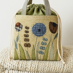 May include: A beige drawstring bag with a floral design. The bag features embroidered flowers in shades of blue, brown, and green. It has a green interior and gray and beige handles. The bag is sitting on a white knitted blanket.