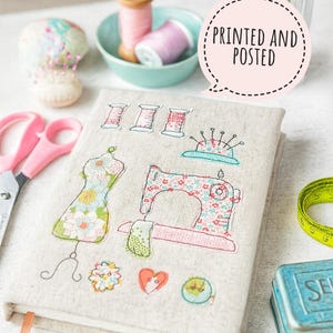 Op de afbeelding: Een beige stoffen journal met geborduurde naaipatronen: een paspop, een naaimachine en garenklosjes. Een roze schaar, een meetlint en een kleine blauwe doos zijn ook aanwezig. De tekst "PRINTED AND POSTED" staat in een tekstballon.