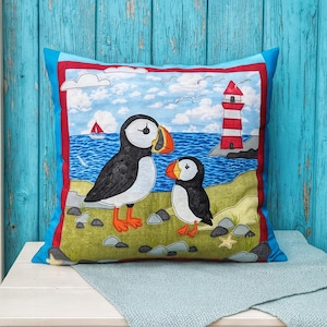Op de afbeelding: Vierkant decoratief kussen met een kleurrijke illustratie van twee papegaaiduikers op een grasrijke oever met een vuurtoren en een zeilboot op de achtergrond. Het kussen heeft een blauwe rand met rode accenten en een blauwe achterkant.