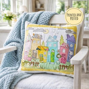Puede incluir: Cojín decorativo bordado con una escena de pueblo colorida. Las casas tienen tejados, puertas y ventanas estampadas, sobre un fondo blanco con un borde amarillo. Texto: "PRINTED AND POSTED" en un bocadillo.