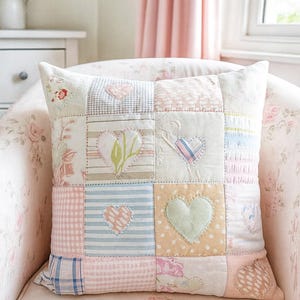 May include: Square patchwork pillow with a variety of pastel fabrics, including floral, striped, and gingham patterns. Each square features a heart applique in various colors. The pillow is placed on a floral patterned chair.