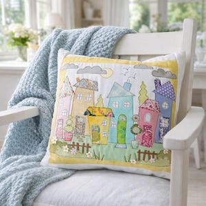 May include: A cushion featuring a hand-stitched village scene with colourful houses, floral accents, and a yellow border. The cushion is placed on a white chair, with a light blue knitted blanket draped over it. The scene includes a picket fence and a small garden.