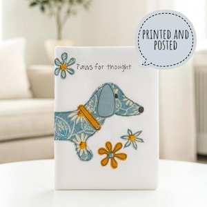 Op de afbeelding: Wit rechthoekig decoratief item met een blauwe bloemen teckel illustratie en de tekst "Paws for thought". De hond heeft een gele halsband. Extra gele en witte bloemen zijn ook te zien. De tekst "PRINTED AND POSTED" staat in een tekstballon.