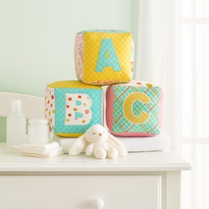 May include: Three colorful fabric alphabet blocks stacked on a white surface. The blocks are yellow, pink, and turquoise, with the letters A, B, and C appliqued on them. A small white stuffed bunny sits in front of the blocks.